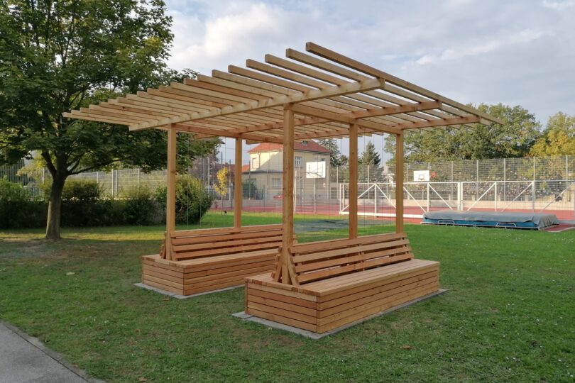 Die Aufnahme zeigt eine neu errichtete Holzpergola mit integrierten Sitzbänken auf einer Rasenfläche im Außenbereich einer Schule. Die Pergola besteht aus hellem, unbehandeltem Holz und bietet Sitzgelegenheiten auf allen vier Seiten. Im Hintergrund ist ein umzäunter Sportplatz mit Basketballkörben und einer Hochsprunganlage zu sehen. Links steht ein großer Laubbaum, rechts und im Hintergrund weitere Bäume und Gebäude. Die Szene wirkt ruhig und gepflegt – vermutlich handelt es sich um eine begrünbare Pergola oder ein Outdoor-Klassenzimmer im Rahmen eines Schulprojekts.