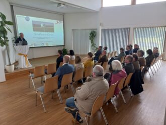 Ein Mann hält einen Vortrag in einem hellen Seminarraum vor etwa 25 sitzenden Zuhörer:innen. Auf der Leinwand wird die Präsentation „SmallWind4Cities“ gezeigt, ein Projekt zu Kleinstwindkraftanlagen für nachhaltige urbane Energiesysteme. Logos von Förderpartnern wie dem Klima- und Energiefonds, der TU Wien und der FFG sind zu sehen. Die Stimmung wirkt aufmerksam und interessiert, große Fenster mit Lamellen lassen Tageslicht in den Raum, Pflanzen sorgen für eine angenehme Atmosphäre.