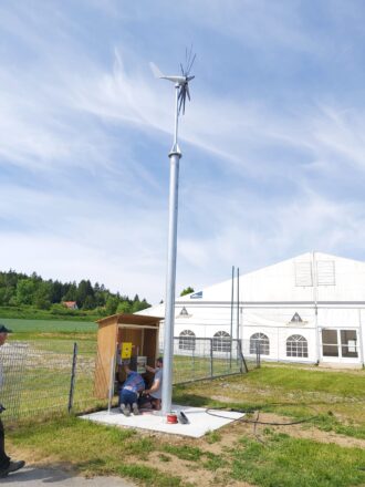 Zwei Personen arbeiten an einem offenen Holzschrank mit elektrischer Ausstattung am Fuß einer kleinen Windkraftanlage. Die Windturbine ist auf einem hohen Metallmast montiert und besitzt einen dreiflügeligen Rotor sowie einen Savonius-Rotor auf der Rückseite. Im Hintergrund stehen ein großes weißes Zelt mit Fenstern und ein grünes Feld mit angrenzendem Wald. Der Himmel ist blau mit leichten Schleierwolken – es herrscht sonniges, ruhiges Wetter. Ein dritter Mann beobachtet die Szene von der Seite.
