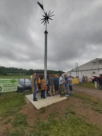 Eine Gruppe von Menschen versammelt sich bei bewölktem Himmel um eine kleine Windkraftanlage, die auf einem hohen Metallmast montiert ist. Die Anlage besteht aus einem weißen dreiflügeligen Rotor vorne und einem schwarzen Savonius-Rotor hinten. Am Boden befindet sich ein kleiner Holzschrank mit Steuerungstechnik, an dem die Teilnehmenden interessiert diskutieren. Im Hintergrund sind ein großes Zelt mit Beschriftung, eine Wiese, ein Werbebanner und weitere Besucher:innen zu sehen. Die Stimmung wirkt neugierig und lehrreich, vermutlich im Rahmen einer Präsentation oder eines Workshops.