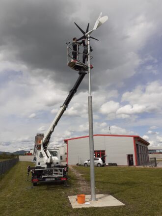 Zwei Techniker stehen in einem Hubsteigerkorb, der an einem weißen Montagefahrzeug befestigt ist, und installieren oder warten eine kleine Windkraftanlage auf einem Metallmast. Die Windturbine hat drei weiße Rotorblätter vorne und einen schwarzen Savonius-Rotor hinten. Das Fahrzeug steht auf einer Wiese neben einem Industriegebäude mit rotem Dachrand. Der Himmel ist bewölkt mit dunklen Wolken im Hintergrund, was auf wechselhaftes Wetter hindeutet.