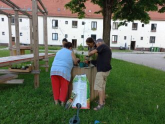 Drei Personen, zwei Frauen und ein Mann, bepflanzen gemeinsam ein Hochbeet auf einer grünen Wiese in einem Wohngebiet. Sie arbeiten konzentriert mit Erde und Pflanzen, ein großer Sack Blumenerde sowie Gartengeräte liegen daneben. Im Hintergrund sind weiße Wohnhäuser mit roten Ziegeldächern und ein Holzgerüst mit Sitzmöglichkeiten zu sehen. Die Szene wirkt gemeinschaftlich und sommerlich.