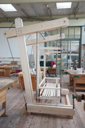 In einer gut ausgestatteten Holzwerkstatt steht ein innovatives, aus hellem Holz gefertigtes Gestell mit drei übereinander hängenden Rahmenstrukturen, die mit Seilen und Metallverbindungen befestigt sind. Die Konstruktion wirkt wie ein modulares Begrünungssystem oder eine bewegliche Rankhilfe, möglicherweise für vertikale Gärten. Rundherum sind klassische Werkbänke, Schraubzwingen und Holzwerkzeuge zu sehen. Die Szenerie deutet auf ein handwerkliches Schulprojekt hin, bei dem das Gestell aktuell gebaut oder getestet wird. Im Hintergrund sind weitere Maschinenräume sichtbar.