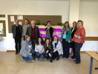 Eine Gruppe von elf Personen steht und kniet in einem Klassenzimmer um eine vertikale Indoor-Begrünungseinheit mit pink-violett leuchtenden Pflanzenlampen. Die Begrünung besteht aus mehreren Ebenen mit eingesetzten Pflanzgefäßen, aus denen grüne Pflanzen wachsen. Die Personen wirken freundlich und posieren für ein Gruppenfoto. Im Hintergrund ist eine Pinnwand mit einigen Zetteln zu sehen, links ein Waschbecken, rechts ein Tisch. Die Atmosphäre ist sachlich, aber positiv – vermutlich handelt es sich um ein schulisches oder forschungsbezogenes Projekt im Bereich Begrünung oder Umweltbildung.