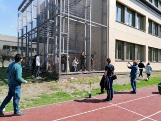 Mehrere Personen, darunter Jugendliche und Erwachsene, befinden sich rund um ein großes, mit Metallgitter gesichertes Treppenhaus an der Außenfassade eines Schulgebäudes. Einige Personen stehen innerhalb des Gitterbereichs, andere beobachten oder fotografieren von außen. Auf dem Boden davor stehen kleine Pflanzen in Töpfen, die auf eine bevorstehende Begrünung hindeuten. Im Vordergrund verläuft eine rote Laufbahn. Die Szene spielt sich bei sonnigem Wetter ab und wirkt wie eine Aktion im Rahmen eines Schulprojekts zur Fassadenbegrünung oder Umweltbildung.