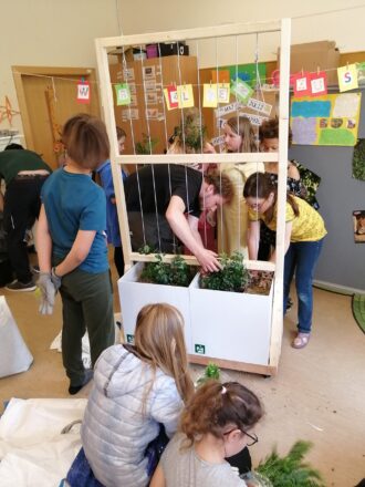 Kinder und ein Erwachsener arbeiten gemeinsam an einem vertikalen Begrünungssystem im Klassenzimmer. Der Aufbau besteht aus einem Holzrahmen mit zwei großen weißen Pflanzgefäßen am Boden, aus denen Kletterpflanzen wachsen sollen. Einige Kinder pflanzen, andere beobachten oder helfen beim Einfüllen der Erde. An der Holzkonstruktion sind beschriftete bunte Kärtchen mit dem Schriftzug „WIR LIEBEN UNSERE SCHULE“ aufgehängt. Im Hintergrund sind Poster, Kalender und weitere kreative Schulmaterialien an der Wand zu sehen. Die Stimmung wirkt lebendig und lehrreich – eine praktische Unterrichtseinheit zum Thema Pflanzen oder Begrünung.