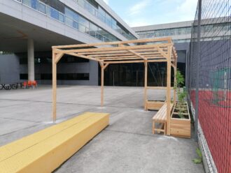 Eine schlichte Holzpergola mit integrierten Sitzbänken und seitlichen Hochbeeten steht auf einem großzügigen, betonierten Schulhof. In den Hochbeeten wachsen junge Pflanzen, die sich an gespannten Seilen emporschlingen sollen. Rechts grenzt ein umzäunter Sportplatz an, links im Hintergrund sind gestapelte orange Stühle und schwarze Tische zu sehen. Die moderne Schularchitektur aus Beton und Glas dominiert den Hintergrund. Die Szene vermittelt ein funktionales, urbanes Schulumfeld mit Bemühungen um Begrünung und Aufenthaltsqualität im Freien.