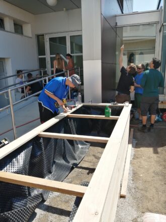 Mehrere Personen arbeiten gemeinsam an einem großen Holzhochbeet im Außenbereich einer Schule. Im Vordergrund schraubt eine Person mit blauem T-Shirt und Sonnenhut an der Holzkonstruktion. Das Hochbeet ist bereits mit schwarzer Noppenfolie ausgekleidet und teilweise mit Erde gefüllt. Im Hintergrund sind weitere Personen bei handwerklichen Tätigkeiten zu sehen, einige stehen an der Fassade und besprechen oder markieren etwas. Andere sitzen auf einer Rampe vor einem Gebäude mit Glastüren. Die Szene wirkt wie ein praxisorientierter Workshop zur Begrünung oder ein schulisches Bauprojekt.