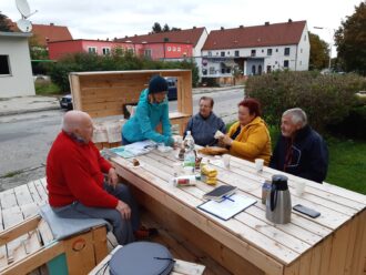 Fünf ältere Menschen sitzen an einem großen Holztisch im Freien und genießen gemeinsam Kaffee und Kuchen. Eine Frau im blauen Mantel und Mütze serviert etwas, während die anderen plaudern und lächeln. Auf dem Tisch stehen Thermoskanne, Getränke, Snacks und ein Notizbuch. Im Hintergrund sind Wohnhäuser mit roten Dächern und ein kleines Geschäft zu sehen. Die Atmosphäre wirkt gesellig und einladend, trotz des kühlen Wetters.