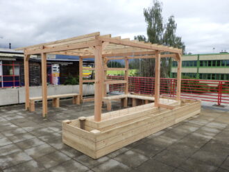 Das Bild zeigt eine hölzerne Pergola-Konstruktion auf einer Dachterrasse oder einem Flachdach eines Schulgebäudes. Die Konstruktion besteht aus naturbelassenem Holz und integriert Hochbeete entlang des äußeren Rahmens. In der Mitte befinden sich Sitzgelegenheiten aus demselben Holzmaterial. Seile oder Drähte sind senkrecht gespannt, vermutlich als Rankhilfe für Kletterpflanzen vorgesehen. Im Hintergrund sind weitere Schulgebäude sowie grüne Bäume sichtbar. Die Szenerie wirkt funktional und auf Begrünung vorbereitet – vermutlich Teil eines Schulprojekts zur Förderung von Outdoor-Lernräumen oder urbanem Gärtnern.