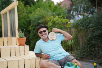 Ein lächelnder Mann mit Sonnenbrille und Hut sitzt entspannt auf einer Holzplattform im Freien. In der Hand hält er ein rundes Holzschild mit der Gravur „Lebe Liebe Lache“. Neben ihm steht ein Tontopf mit einer Pflanze. Im Vordergrund sind Flaschen und eine grüne Tasche zu sehen, im Hintergrund dichte grüne Vegetation. Die Stimmung ist fröhlich und locker, das Wetter wirkt sommerlich warm.