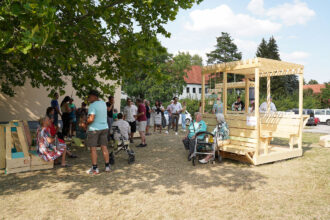 Eine Gruppe von Menschen unterschiedlichen Alters versammelt sich an einem sonnigen Tag unter einem großen Baum auf einer trockenen Wiese. Links sitzen und stehen ältere Personen, teils mit Rollatoren, auf bunten Holzsitzmöbeln. Rechts ist ein großes, überdachtes Holzmodul mit Sitzbänken und Spielwand aufgebaut, davor unterhalten sich zwei ältere Frauen. Im Hintergrund stehen weitere Personen in Grüppchen beisammen und plaudern. Die Atmosphäre wirkt lebendig, gesellig und generationenübergreifend.