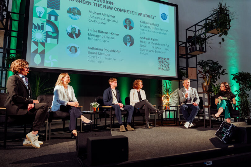 Podiumsdiskussion mit sechs Personen auf der Bühne vor einer Präsentationsfolie zum Thema 'Is Green the New Competitive Edge?'