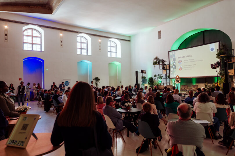 Großer Saal mit Publikum, das einer Präsentation auf einer Bühne mit Leinwand folgt, beleuchtet mit buntem Licht