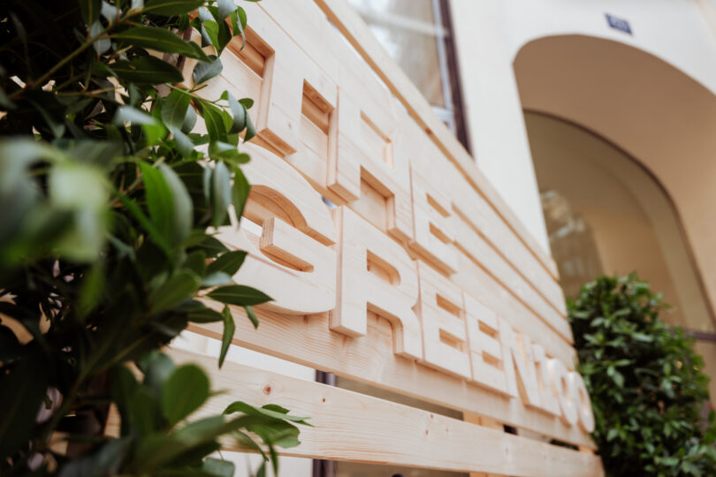 Holzschild mit weißen Buchstaben 'THE GREEN' vor einem Gebäude mit Bogen und grünen Pflanzen