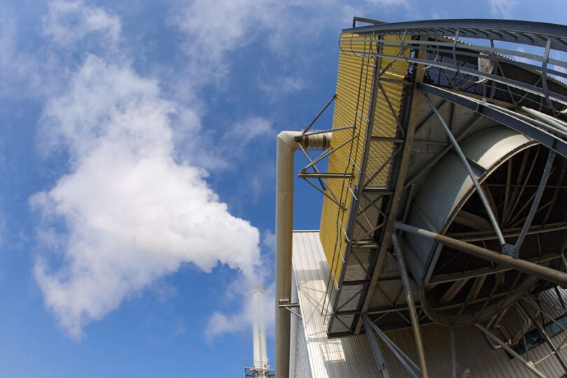Hoher Industrieschornstein stößt weißen Dampf in den Himmel aus. Im Vordergrund ist ein moderner Gebäudeteil mit Metallerüst zu sehen, der Schornstein steht vor einem blauen Himmel mit leichten Wolken.