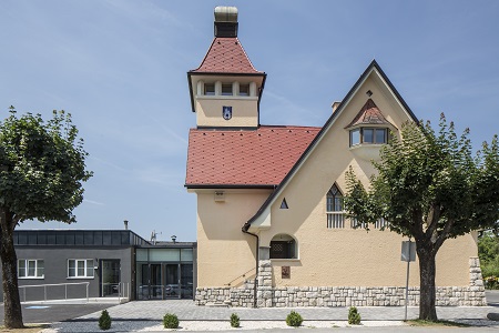Helles Gebäude mit rotem Satteldach und Turm, kombiniert mit modernem Anbau, davor Bäume und gepflasterter Gehweg.