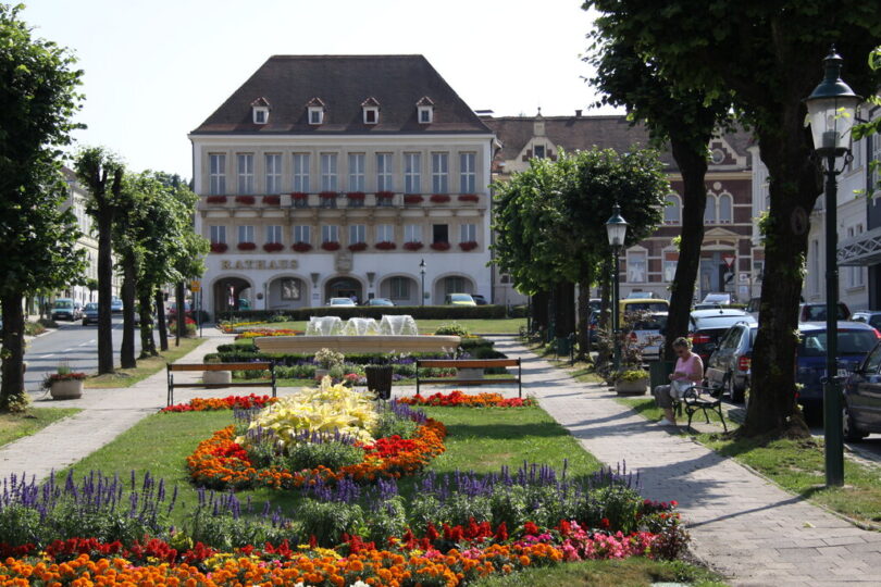 Eine gepflegte Parkanlage mit bunten Blumenbeeten, Sitzbänken und einem Brunnen führt zu einem historischen Rathausgebäude mit roten Fensterblumen. Rechts sitzt eine Frau auf einer Bank im Schatten von Bäumen. Autos parken entlang der Straße, und im Hintergrund sind weitere Altbauten zu sehen.