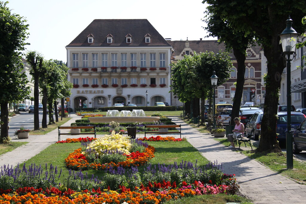 Eine gepflegte Parkanlage mit bunten Blumenbeeten, Sitzbänken und einem Brunnen führt zu einem historischen Rathausgebäude mit roten Fensterblumen. Rechts sitzt eine Frau auf einer Bank im Schatten von Bäumen. Autos parken entlang der Straße, und im Hintergrund sind weitere Altbauten zu sehen.