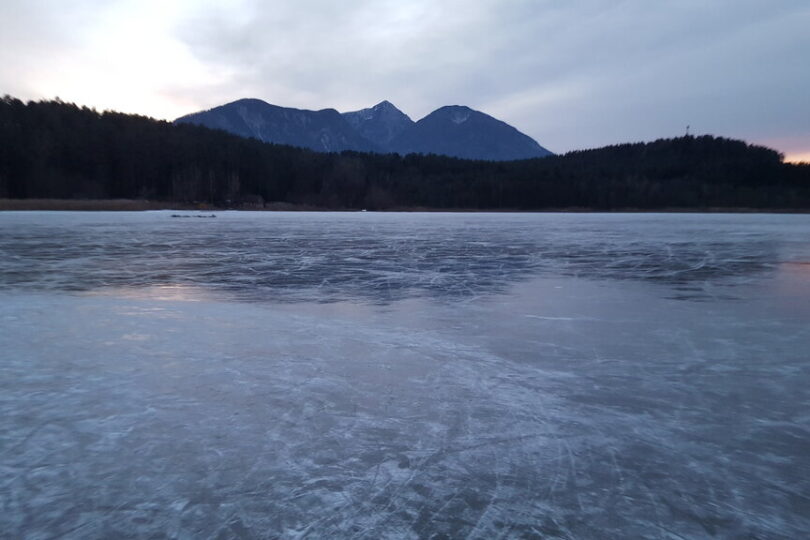 Zu einer winterlichen Abendstimmung gefrorener See mit glatter Eisoberfläche, umgeben von dunklem Wald. Im Hintergrund erheben sich schneebedeckte Berge unter einem bewölkten Himmel.