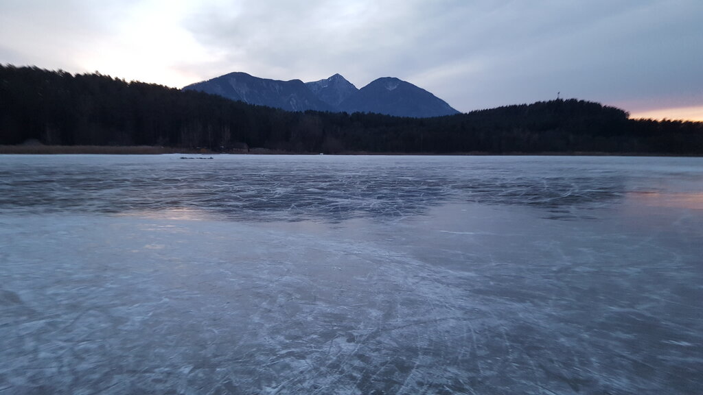 Zu einer winterlichen Abendstimmung gefrorener See mit glatter Eisoberfläche, umgeben von dunklem Wald. Im Hintergrund erheben sich schneebedeckte Berge unter einem bewölkten Himmel.