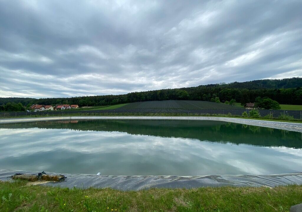 Ein künstlich angelegtes Wasserbecken in ländlicher Umgebung, umgeben von Wiesen, einem mit Folie abgedeckten Hang (vermutlich für Landwirtschaft), einem Wald im Hintergrund und einem Dorf mit roten Dächern am linken Bildrand unter einem bewölkten Himmel.