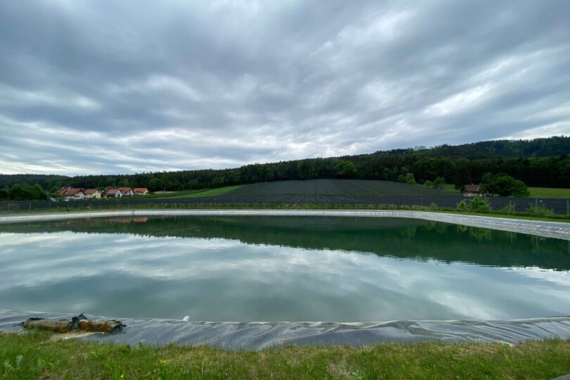 Ein künstlich angelegtes Wasserbecken in ländlicher Umgebung, umgeben von Wiesen, einem mit Folie abgedeckten Hang (vermutlich für Landwirtschaft), einem Wald im Hintergrund und einem Dorf mit roten Dächern am linken Bildrand unter einem bewölkten Himmel.