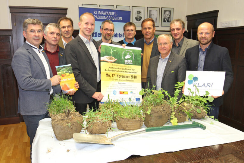 Eine Gruppe von Männern steht in einem Veranstaltungsraum hinter einem Tisch, auf dem Erdklumpen mit Pflanzen und ein Spaten liegen. Sie halten verschiedene Plakate und Broschüren zum Thema Boden, Humusaufbau und Klimawandelanpassung. Im Hintergrund sind Banner mit der Aufschrift „Klimawandel-Anpassungsregion“ sowie Porträtfotos an der Wand zu sehen.