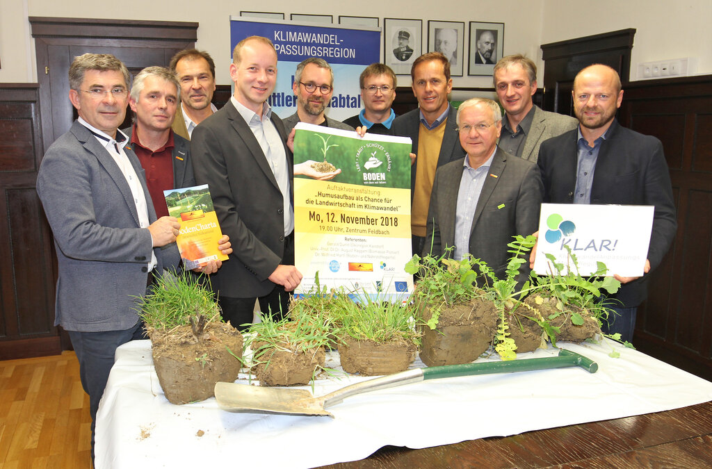 Eine Gruppe von Männern steht in einem Veranstaltungsraum hinter einem Tisch, auf dem Erdklumpen mit Pflanzen und ein Spaten liegen. Sie halten verschiedene Plakate und Broschüren zum Thema Boden, Humusaufbau und Klimawandelanpassung. Im Hintergrund sind Banner mit der Aufschrift „Klimawandel-Anpassungsregion“ sowie Porträtfotos an der Wand zu sehen.
