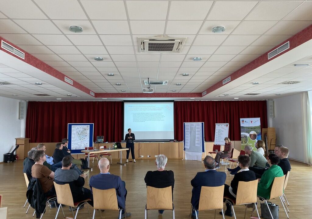 Gruppensituation in einem Konferenzraum: etwa 20 Personen sitzen im Stuhlkreis, vorne präsentiert eine Person an einer Leinwand. Im Raum stehen Karten und Flipcharts. Atmosphäre wirkt partizipativ, vermutlich Workshop oder Planungstreffen zu regionalen Themen.