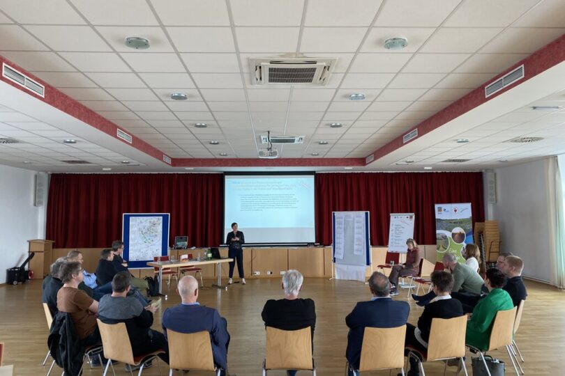 Gruppensituation in einem Konferenzraum: etwa 20 Personen sitzen im Stuhlkreis, vorne präsentiert eine Person an einer Leinwand. Im Raum stehen Karten und Flipcharts. Atmosphäre wirkt partizipativ, vermutlich Workshop oder Planungstreffen zu regionalen Themen.