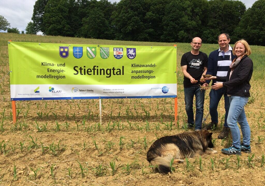 Drei Personen stehen auf einem Acker vor einem großen grünen Banner mit der Aufschrift „Stiefingtal – Klima- und Energiemodellregion / Klimawandelanpassungsmodellregion“. Zwei von ihnen halten Erdklumpen mit Pflanzen in den Händen. Vor ihnen liegt ein großer Hund im Gras. Im Hintergrund sind Bäume und blauer Himmel zu sehen.