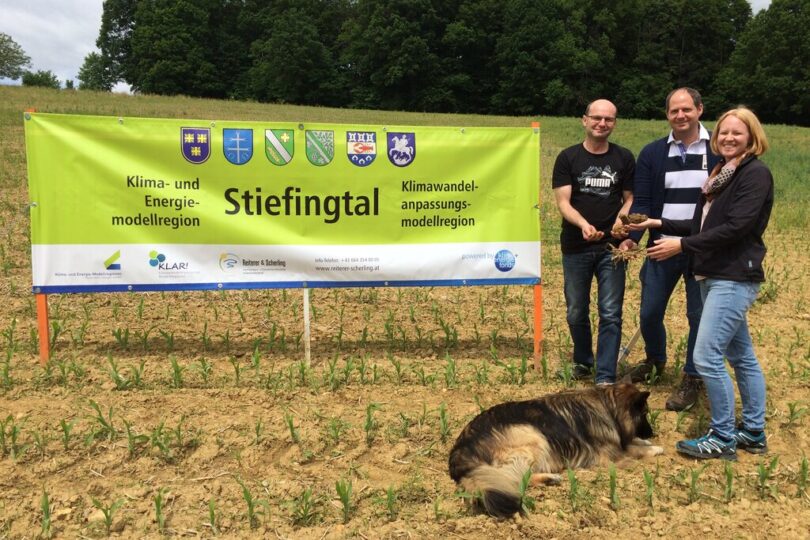 Drei Personen stehen auf einem Acker vor einem großen grünen Banner mit der Aufschrift „Stiefingtal – Klima- und Energiemodellregion / Klimawandelanpassungsmodellregion“. Zwei von ihnen halten Erdklumpen mit Pflanzen in den Händen. Vor ihnen liegt ein großer Hund im Gras. Im Hintergrund sind Bäume und blauer Himmel zu sehen.