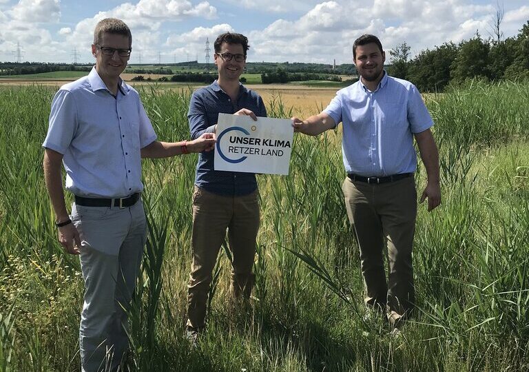 Drei Männer stehen auf einer Wiese mit hohem Gras und halten gemeinsam ein Schild mit der Aufschrift „Unser Klima Retzer Land“. Im Hintergrund sind Felder, Bäume und ein leicht bewölkter Himmel zu sehen.