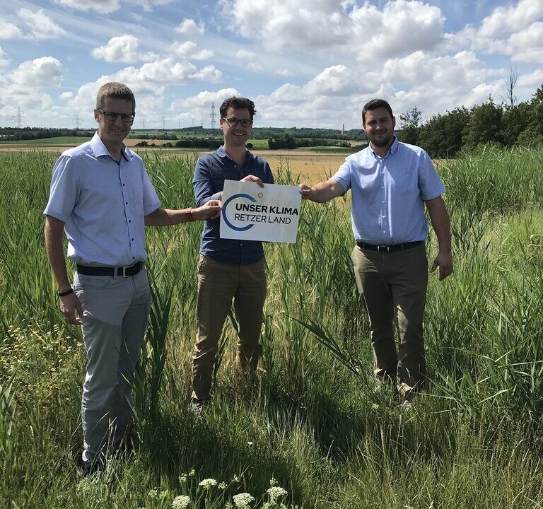 Drei Männer stehen auf einer Wiese mit hohem Gras und halten gemeinsam ein Schild mit der Aufschrift „Unser Klima Retzer Land“. Im Hintergrund sind Felder, Bäume und ein leicht bewölkter Himmel zu sehen.