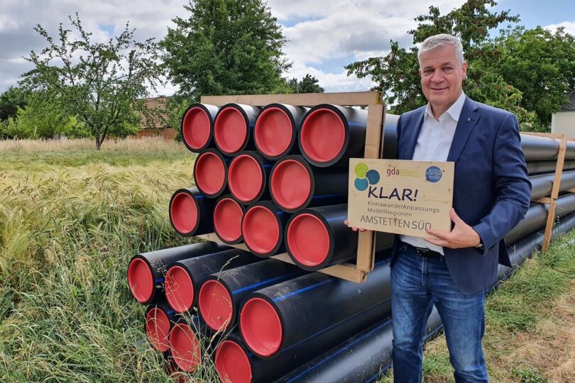 Ein Mann in Anzug steht auf einem Feld neben gestapelten schwarzen Kunststoffrohren mit roten Endkappen und hält ein Schild mit der Aufschrift „KLAR! KlimawandelAnpassungs Modellregionen – Amstetten Süd“. Im Hintergrund sind Bäume, eine Wiese und ein Haus zu sehen.