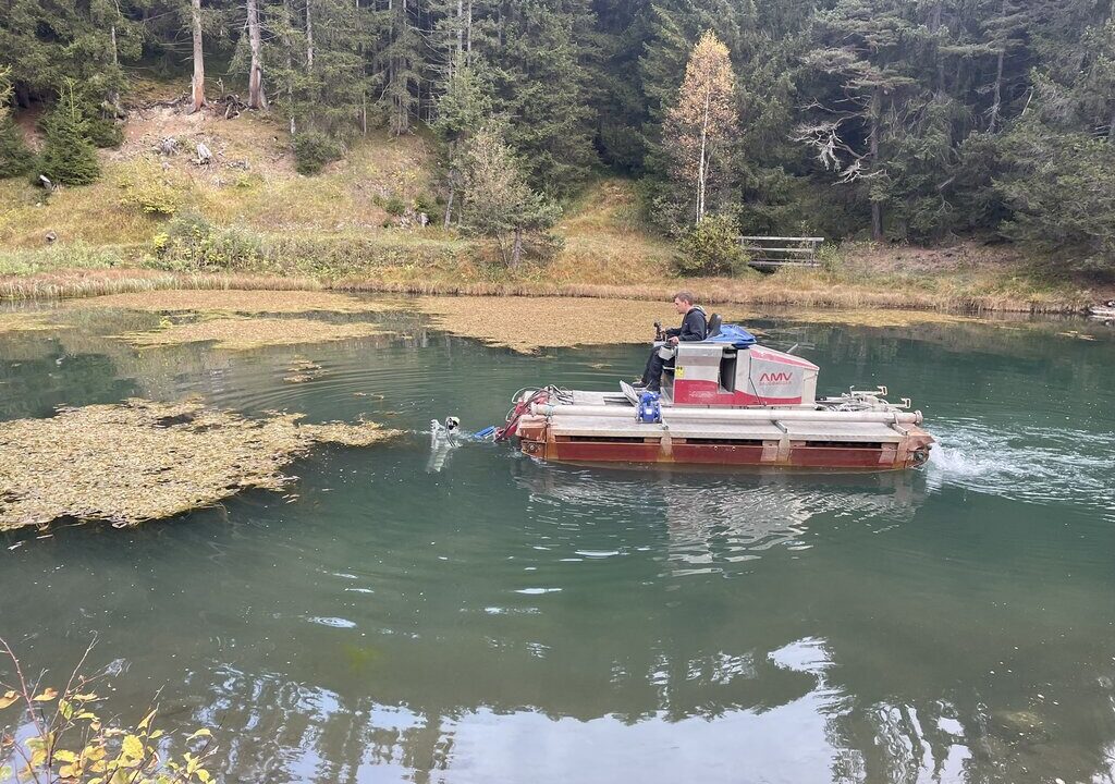 Kleines Spezialboot mit Saugbagger wird auf einem bewaldeten Teich eingesetzt, um Wasserpflanzen oder Sedimente zu entfernen. Ein Mann steuert das Gerät, das gezielt schwimmende Pflanzenmatten ansaugt. Im Hintergrund dichter Nadelwald.