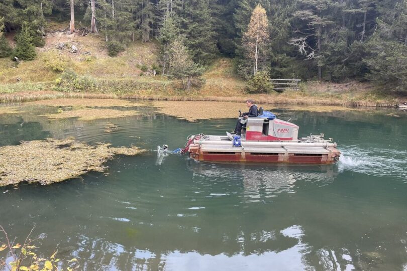 Kleines Spezialboot mit Saugbagger wird auf einem bewaldeten Teich eingesetzt, um Wasserpflanzen oder Sedimente zu entfernen. Ein Mann steuert das Gerät, das gezielt schwimmende Pflanzenmatten ansaugt. Im Hintergrund dichter Nadelwald.