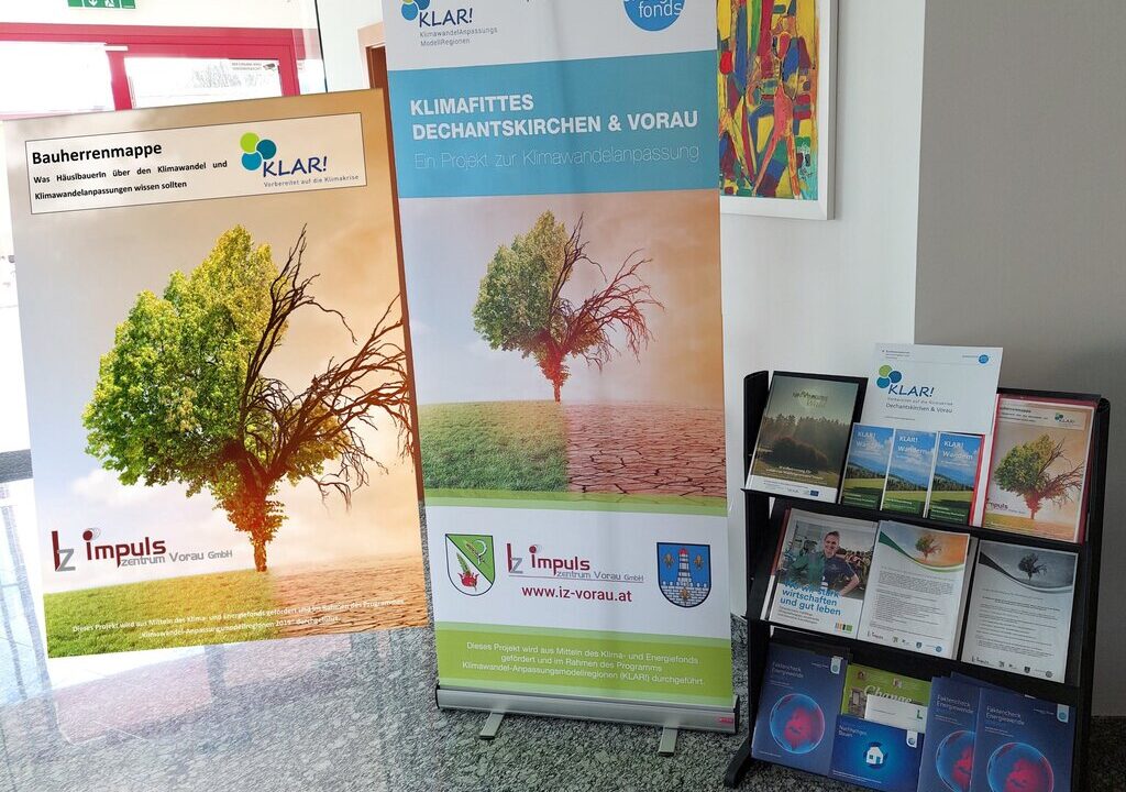 Informationsstand zur Klimawandelanpassung in einem modernen Gebäude. Zwei Roll-Up-Banner mit dem Bild eines Baums, der zur Hälfte grün und zur Hälfte verdorrt ist, stehen nebeneinander. Der rechte Banner trägt die Aufschrift „Klimafittes Dechantskirchen & Vorau“. Daneben befindet sich ein schwarzes Regal mit Broschüren, Flyern und Informationsmaterialien zum Thema Klimawandel und regionaler Anpassung.