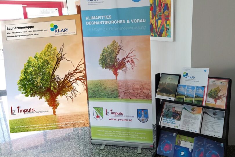 Informationsstand zur Klimawandelanpassung in einem modernen Gebäude. Zwei Roll-Up-Banner mit dem Bild eines Baums, der zur Hälfte grün und zur Hälfte verdorrt ist, stehen nebeneinander. Der rechte Banner trägt die Aufschrift „Klimafittes Dechantskirchen & Vorau“. Daneben befindet sich ein schwarzes Regal mit Broschüren, Flyern und Informationsmaterialien zum Thema Klimawandel und regionaler Anpassung.