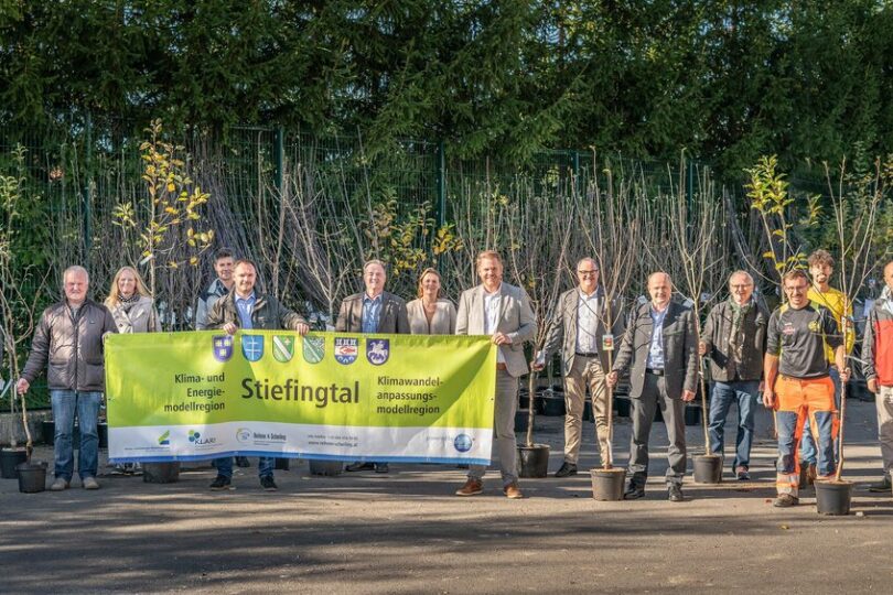 Gruppe von etwa 15 Personen posiert im Freien vor einer Reihe junger Bäume in Töpfen. In der Mitte halten sie ein großes Banner der Klima- und Energiemodellregion Stiefingtal. Die Aktion steht sichtbar im Zeichen klimafreundlicher Begrünung.