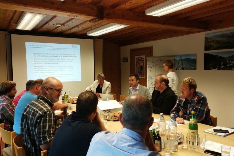 Eine Gruppe von Menschen sitzt in einem Holzkonferenzraum um einen Tisch und diskutiert. Auf dem Tisch stehen Getränke und Unterlagen. Im Hintergrund ist eine Präsentation mit dem Titel „Lernen von der Katastrophe“ zu sehen. An der Wand hängen Poster mit Informationen zur Klimawandelanpassung in der Region Ennstal.