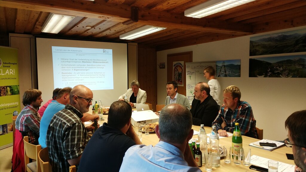 Eine Gruppe von Menschen sitzt in einem Holzkonferenzraum um einen Tisch und diskutiert. Auf dem Tisch stehen Getränke und Unterlagen. Im Hintergrund ist eine Präsentation mit dem Titel „Lernen von der Katastrophe“ zu sehen. An der Wand hängen Poster mit Informationen zur Klimawandelanpassung in der Region Ennstal.