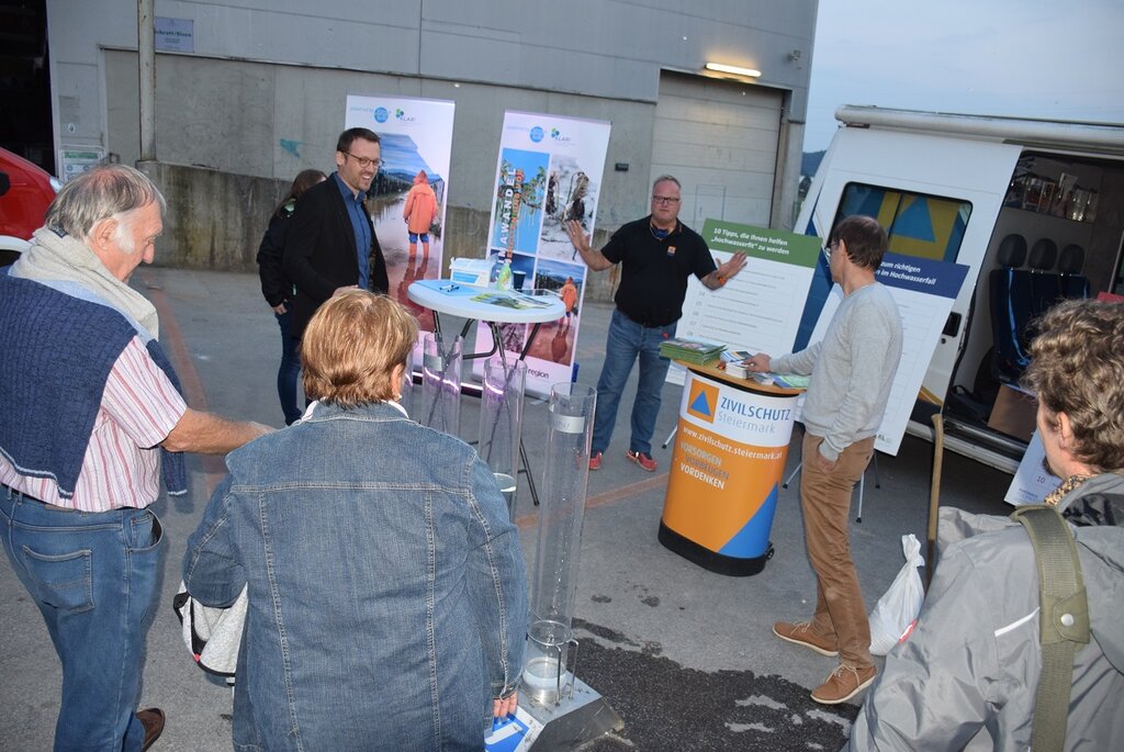 Eine Gruppe von Menschen steht bei einer Informationsveranstaltung im Freien vor einem mobilen Infostand des Zivilschutzes Steiermark. Ein Mann erklärt etwas anhand von Infotafeln zum Thema Hochwasserschutz und Klimawandelanpassung. Im Hintergrund ist ein Fahrzeug mit geöffneter Tür und Plakaten zu sehen.