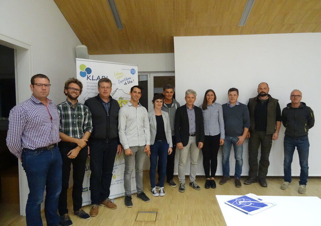 Eine Gruppe von zwölf Personen steht in einem modernen Raum mit Holzdecke und hellem Boden. Im Hintergrund befindet sich ein Roll-up-Banner mit dem KLAR!-Logo (Klimawandel-Anpassungsmodellregion Pongau). Die Personen posieren für ein Gruppenfoto, vermutlich im Rahmen eines Workshops oder einer Informationsveranstaltung zur Klimaanpassung. Auf dem Tisch im Vordergrund liegt ein blauer Notizblock.