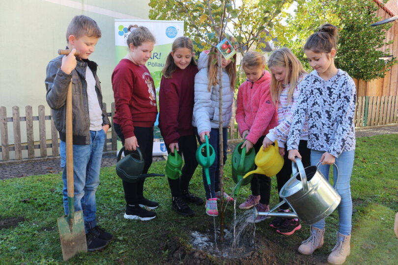 Eine Gruppe von sieben Kindern steht auf einer Wiese und gießt gemeinsam einen frisch gepflanzten jungen Baum mit Gießkannen. Ein Junge hält zusätzlich einen Spaten. Im Hintergrund sind ein Holzzaun, ein Gebäude und ein Baum sowie ein Banner mit dem Logo der KLAR!-Initiative zu sehen.