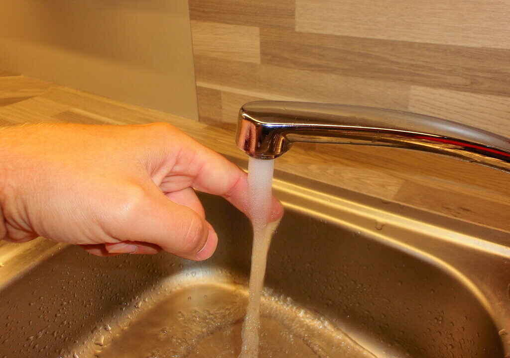 Das Bild zeigt eine Nahaufnahme eines Wasserhahns über einem Spülbecken. Aus dem Hahn fließt ein klarer Wasserstrahl. Eine Hand testet die Wassertemperatur oder den Wasserfluss mit dem Daumen. Im Hintergrund ist eine Küchenrückwand aus Holzpaneelen sichtbar.
