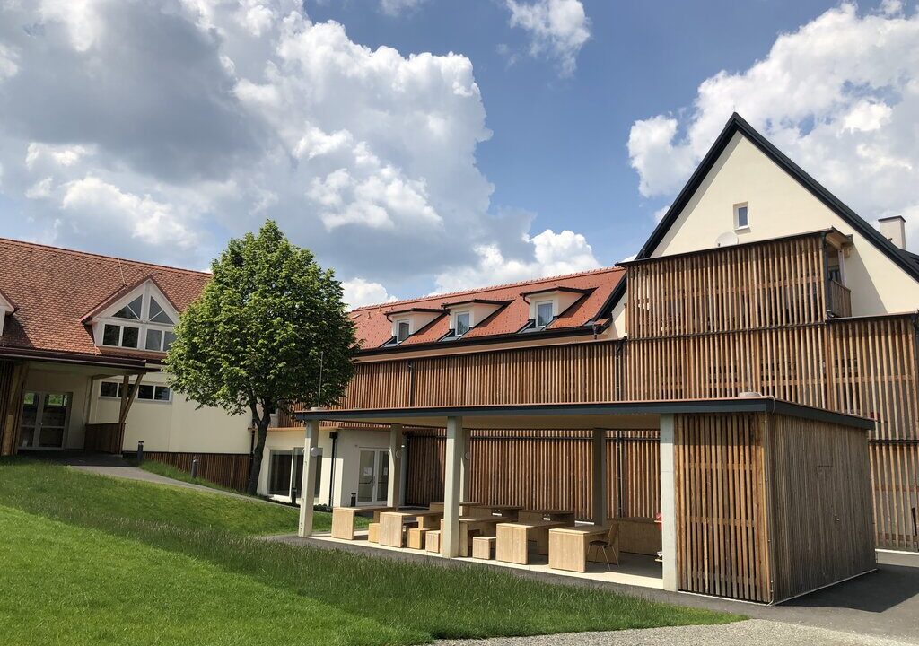 Ein modernes Schulgebäude mit Holzverschalung und roten Ziegeldächern bei sonnigem Wetter. Im Vordergrund befindet sich eine überdachte Sitzfläche mit Holzbänken und -tischen sowie eine gepflegte Rasenfläche und ein einzelner Baum. Der Himmel ist teils bewölkt.