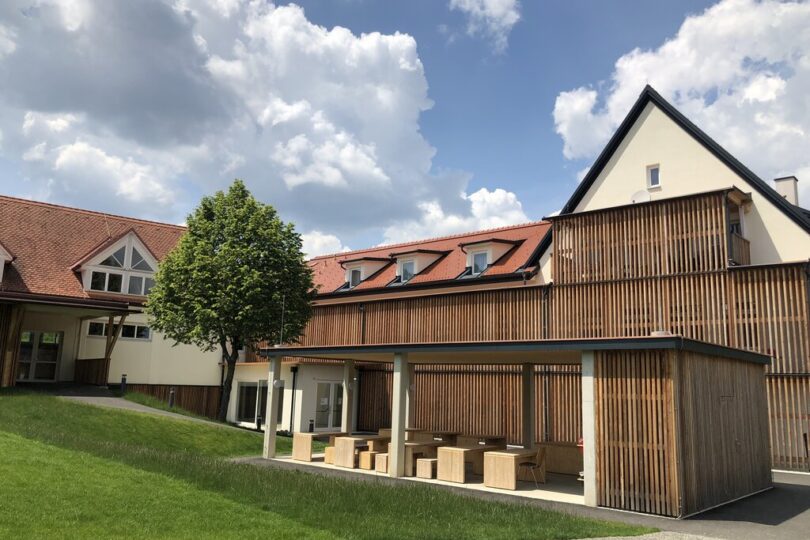 Ein modernes Schulgebäude mit Holzverschalung und roten Ziegeldächern bei sonnigem Wetter. Im Vordergrund befindet sich eine überdachte Sitzfläche mit Holzbänken und -tischen sowie eine gepflegte Rasenfläche und ein einzelner Baum. Der Himmel ist teils bewölkt.