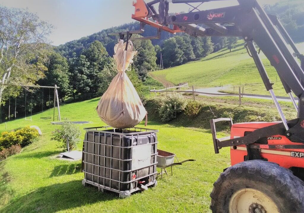 Ein Traktor mit Frontlader hebt einen großen, in einem Tuch verpackten Ballen über einen IBC-Tank. Die Szene spielt sich auf einer Wiese in hügeliger Landschaft ab. Im Hintergrund sind Bäume, ein Spielplatz und ein Feldweg sichtbar.